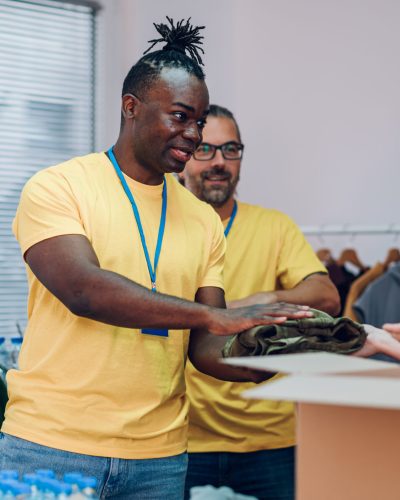 Diverse volunteers wearing yellow t shirt at donations stand separating donation stuff and putting them in a cardboard boxes. Concept of a charitable foundation. Focus on an african american man.